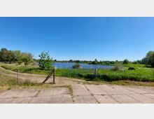 Eine ländliche Landschaft mit einem Fluss oder See im Hintergrund, umgeben von grüner Wiese und Bäumen. Im Vordergrund befinden sich ein unbefestigter Weg, ein Tor sowie ein Zaun.