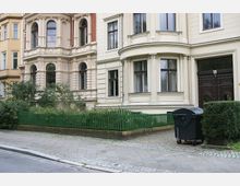 Fassadenansicht eines historischen Wohnhauses mit verzierten Fensterrahmen und heller Putzfassade. Im Vordergrund ein grüner Zaun, kleinere Sträucher, ein gepflasterter Gehweg und eine schwarze Mülltonne.