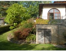 Ein Garten mit einer Rasenfläche und verschiedenen Sträuchern, darunter ein großer grüner Baum. Im Hintergrund befindet sich ein Gebäude mit heller Fassade, einem großen, runden Fenster und einer Veranda mit Metallgeländer. Im unteren Bereich der Hauswand ist eine rustikale Holztür in einer Steinfassade sichtbar.