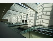 Modernes Innenraumdesign mit einer großen Halle und einem Oberlicht, das geometrische Schatten auf die hellen Betonwände wirft. Der Bereich verfügt über Glasgeländer, eine Rolltreppe und Sitzgelegenheiten auf einer erhöhten Plattform.