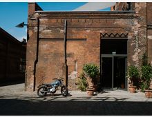 Exterior of an old red-brick industrial building with weathered masonry, exposed pipes and wiring, and a large glass doorway opening onto a cobblestone courtyard. Potted shrubs line the entrance, and a motorcycle is parked against the brick wall under bright daylight.