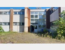 Flaches, rechteckiges Gebäude im Stil der 1970er Jahre mit großflächigen Fenstern und Putzfassade, teilweise verwittert. Der Vorplatz ist mit Steinplatten bedeckt und von unkontrolliert wachsenden Pflanzen überwuchert.