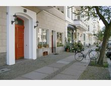 Straßenszene mit Altbau-Häusern, Geschäften und Balkonen; im Vordergrund ein rotes Holztor sowie ein Gehsteig mit Pflastersteinen, einem geparkten Fahrrad und Pflanzen auf dem Bürgersteig. Links steht ein Baum, rechts sind weitere Gebäude und parkende Autos sichtbar.