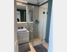 Modernes Badezimmer mit Glas-Duschkabine, Regendusche und beigefarbenen Fliesen. Daneben ein weißer Waschtisch mit Spiegel und darüber integrierter Beleuchtung; der Boden ist mit zwei grauen Teppichen ausgelegt.