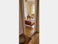 Blick in ein schlicht eingerichtetes Zimmer mit Holzboden, gelben Vorhängen, einem kleinen Holztisch, zwei Stühlen und einem Sofa. Durch das Fenster ist ein Teil eines Gebäudes im Hintergrund erkennbar.