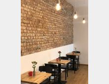 Ein kleines Café mit Tischen aus hellem Holz, schwarzen Stühlen und einer Wand mit freigelegtem Backstein. Jeder Tisch ist mit einer Blumenvase und einer farbigen Tasse dekoriert, während von der Decke einfache Glühbirnen hängen.