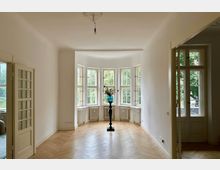 Heller Wohnraum mit einem Erker aus großen Sprossenfenstern, darunter weiße Heizkörper, und einem Parkettboden in Fischgrätmuster. Im Erker steht eine Vase mit Blumen auf einem hohen, dunklen Sockel; im Vordergrund sind geöffnete Doppeltüren mit Glaseinsätzen.