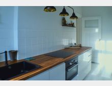 Moderne Küche mit weißen Fliesen an der Wand, einer Arbeitsplatte aus Holz, elektrischem Kochfeld, integriertem Backofen und einem schwarzen Spülbecken. Drei schwarze Hängelampen beleuchten den Raum, und an der Wand befindet sich ein kleines Regal mit Gewürzen und Ölen.