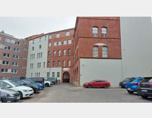 Ziegelgebäude mit modernen Fenstern und Bogeneingang, umgeben von einem Hof mit geparkten Autos. Das Gebäude kombiniert Backstein- und Putzfassaden und wirkt wie ein renoviertes Industrie- oder Bürogebäude.