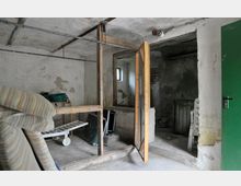 Kellerraum mit einer ungestrichenen Betondecke und abgenutzten Wänden, ausgestattet mit einem Holzgitter und Drahtzaun als Abtrennung. Im Raum befinden sich Gegenstände wie ein zusammengeklappter Liegestuhl, gestapelte Gartenmöbel und ein grünes Tor.