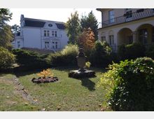 Ein gepflegter Garten mit einer steinernen Skulptur in der Mitte, umgeben von Sträuchern und herbstlichen Pflanzen; im Hintergrund befinden sich ein weißes Gebäude mit verziertem Giebel und ein helles Gebäude mit Arkadengang.