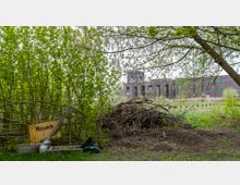 Ein bewaldetes Gelände mit einem gelben Ortsschild „Nauen“, das an einem provisorischen Weidenzaun lehnt. Im Hintergrund ist ein größeres, dunkelbraunes Backsteingebäude mit hohen Fenstern und grünen Wiesen im Vordergrund zu sehen.