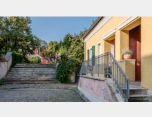 Eine gelb gestrichene Hausfassade mit grünen Fensterläden und einem Geländer führt zu einer Treppe. Im Hintergrund ist eine steinerne Treppe von Pflanzen umgeben, die zu einem rosa Gebäude und einem baumreichen Bereich führt.