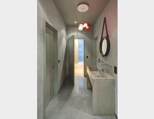 Ein schmaler Flur mit glatten grauen Wänden und glänzendem Steinboden. Auf der rechten Seite ein längliches Waschbecken aus Beton mit zwei Wasserhähnen und einem runden Spiegel darüber, sowie eine bunte Hängelampe an der Decke; im Hintergrund eine geöffnete Tür, die zu einem Raum mit Holzboden führt.