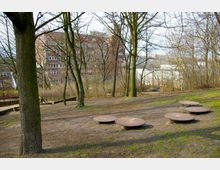 Ein kleiner Park mit mehreren Bäumen ohne Laub, vereinzelten Grasflächen und federnden Sitzscheiben auf dem Boden. Im Hintergrund sind mehrstöckige Backsteingebäude und weitere städtische Strukturen zu sehen.
