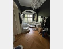 Großes Badezimmer mit dunklen Wänden, einem Hartholzboden im Fischgrätmuster und einem Kronleuchter an der Decke. Der Raum verfügt über eine bodentiefe Dusche, ein großes Fenster mit Rundbogen, zwei Waschbecken mit Spiegeln und einen Bereich mit Badewanne sowie Toiletten.