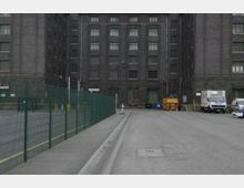 Großes, mehrstöckiges Gebäude aus dunklem Backstein mit zahlreichen Fenstern, umgeben von einem gepflasterten Hof. Im Vordergrund steht ein grüner Metallzaun, während rechts geparkte Fahrzeuge, Müllcontainer und Baumaschinen sichtbar sind.