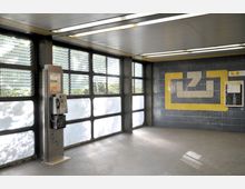 Innenraum einer Haltestelle mit schlichtem Design, gekennzeichnet durch große Fenster mit vergitterten Elementen. An der Wand rechts befindet sich ein gelbes Symbol auf blauen Fliesen, daneben eine Informationstafel, während links ein öffentliches Münztelefon steht.
