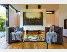 Moderner Wohnraum mit Holzfußboden, zwei schwarzen Ledersesseln und einem niedrigen Glastisch mit Zeitschriften. Große bodentiefe Fenster und eine zentrale Wand mit großformatigem Bild prägen den Raum, daneben ist ein eingebauter Kamin zu sehen.