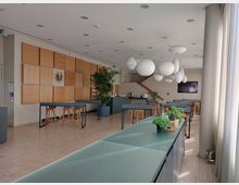 Moderner Gemeinschaftsraum mit hellen Wänden, großen Fensterfronten und hängenden weißen Lampenschirmen. Der Raum ist mit hohen Tischen, Barhockern, einer bepflanzten Theke und einem dekorativen Holzpaneel an der Wand gestaltet.