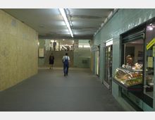 Unterirdischer Fußgängerdurchgang mit grünen Fliesenwänden, einer kleinen Bäckerei auf der rechten Seite und einer Treppe, die zu einer U-Bahn-Station führt. Beleuchtung erfolgt durch lange Deckenleuchten, und auf der linken Seite sind Sperrholzwände mit Graffiti zu sehen.