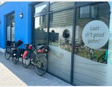 Fahrräder mit Gepäcktaschen stehen an der gläsernen Front eines Gebäudes mit blauer Fassade. Auf der Scheibe ist eine Sprechblase mit dem Text "Laat di’t good gahn!" angebracht.