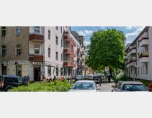 Straßenszene in einem Wohnviertel mit mehrstöckigen Gebäuden und Balkonen, links eine Kneipe mit Sitzmöglichkeiten im Außenbereich. Rechts ein großer Baum, Autos parken entlang der Straße, und ein Schild zeigt eine Tempo-30-Zone an.