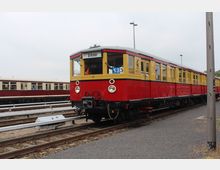Ein historischer Zug in rot-gelber Lackierung steht auf Gleisen in einem Bahnhofsbereich. Die Zuggarnitur trägt die Linienanzeige „S33“ und das Ziel „Erkner“, im Hintergrund sind weitere Züge zu sehen.