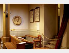 Innenansicht eines Kirchenraums mit einer Holzbank und einer Orgelkonsole im Vordergrund. An der Wand ein Rundrelief, das ein Gesicht darstellt, sowie zwei hölzerne Tafeln mit Text; rechts eine Treppe, teilweise von einem roten Vorhang verdeckt.