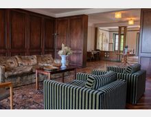 Wohnzimmer mit dunkler Holzvertäfelung an den Wänden, gepolsterten Möbeln mit gemusterten Stoffen, einem orientalischen Teppich auf Holzboden und einem kleinen Holztisch mit Blumenvase. Im Hintergrund ein angrenzender Raum mit großen Fenstern und klassischen Holzmöbeln.