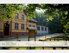 Gelbes Gebäude mit roten Backsteinverzierungen, beschriftet mit „Zur alten Eiche“, Teil einer Waldgaststätte. Vor dem Gebäude stehen Holzbänke und ein Informationsschild, umgeben von Bäumen und Waldlandschaft.