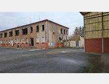 Verlassener Industriehof mit einem zweigeschossigen, teils verputzten Backsteingebäude mit zerbrochenen Fenstern und zugemauerten Öffnungen sowie einem Eingang mit Stufen. Rechts steht eine große Hallenwand aus Wellplatten und rotem Mauerwerk, davor eine breite asphaltierte Freifläche.