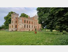 Verlassene, zweistöckige Backsteinvilla mit starken Schäden an der Fassade, flankiert von einem kleinen Turm. Die Villa steht in einer grünen Wiese, umgeben von Bäumen, wobei der Himmel leicht bewölkt ist.