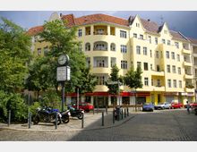 Gelbes, mehrstöckiges Wohngebäude mit hellrotem Ziegeldach und Balkonen, umgeben von Bäumen und einem gepflasterten Platz. Im Vordergrund stehen eine runde Uhr auf einem Mast, Straßenschilder („Böhmischer Platz“) sowie geparkte Motorräder und Autos.