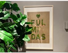 Ein Wandbild mit weißem Rahmen, das den Schriftzug „FULMIDAS“ aus grünem Material zeigt, zusammen mit einem herzförmigen Symbol darüber. Links im Bild steht eine große Zimmerpflanze mit glänzend grünen Blättern, vor einer hellgrauen Wand.