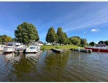 Ein kleiner Hafen mit mehreren Booten, die an Holzstegen festgemacht sind. Im Hintergrund befinden sich ein grasbewachsener Uferbereich mit hohen Bäumen und Gebäuden unter einem klaren blauen Himmel.