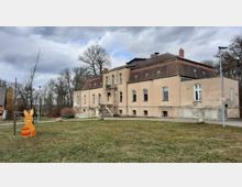 Historisches Gebäude mit beiger Fassade und rotem Ziegeldach, umgeben von Bäumen und einer Rasenfläche. Im Vordergrund steht eine bunte Holzfigur in Form eines Hasen mit Ostermotiven.