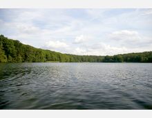 Weitläufiger See umgeben von dicht bewaldeten Ufern, unter einem leicht bewölkten Himmel. Die Wasseroberfläche zeigt leichte Wellen.
