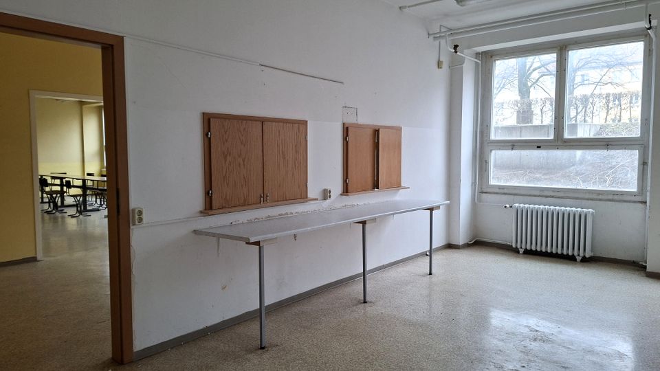 Leerer Raum in einem älteren Schul- oder Bürogebäude mit weiß gestrichenen Wänden, großem Fenster und Heizkörper darunter. An der Wand befindet sich eine lange Ablagefläche mit zwei Holzklappen-Fensternischen; links führt eine offene Tür in einen weiteren Raum mit Tischen.