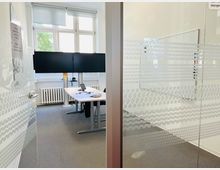 Ein moderner Büroraum mit Glaswänden, teilweise mit Milchglas-Dekor. Im Inneren steht ein Tisch mit einem Laptop, Kabeln und zwei großen Monitoren, ergänzt durch ein Fenster mit Blick auf grüne Bäume und eine weiße Heizung darunter.