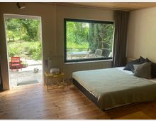 Schlafzimmer mit Holzfußboden und Holzdecke, einem Doppelbett neben einem großen Panoramafenster mit Vorhang. Links führt eine offene Tür auf eine Terrasse und in einen grünen Garten; draußen sind Sitzmöbel sichtbar.