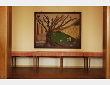 Ein einfacher Raum mit Holzboden, hellbeigen Wänden und einem großen Wandteppich, der eine Landschaft mit einem Baum, einer Wiese und Figuren darstellt. Entlang der Wand steht ein langer Tisch mit rotem, gemustertem Tuch.