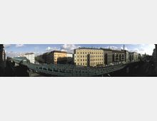 Panoramablick auf eine städtische Straße mit gelben und hellen Wohngebäuden im klassischen Berliner Altbaustil. Im Vordergrund verläuft eine erhöhte U-Bahn-Trasse mit einer Station, und die Straße darunter ist von Fahrzeugen und Fußgängern belebt.