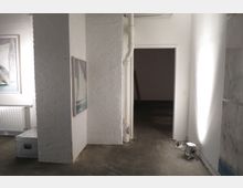 Ein minimalistischer Ausstellungsraum mit weißen Backsteinwänden, einem Betonboden und Kunstwerken an den Wänden. Im Hintergrund führt eine Tür in einen dunkleren Raum; ein Strahler steht auf dem Boden an der rechten Wand.