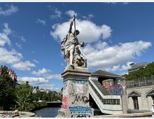 Statue einer historischen Figur, die einen Speer hält, auf einem grauen, mit Graffiti bedeckten Sockel, daneben ein Treppenaufgang mit moderner Architektur. Der Hintergrund zeigt einen Fluss, Gebäude und Bäume unter einem sonnigen Himmel mit wenigen Wolken.