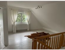 Helles Zimmer mit Dachschräge, weiß gestrichenen Wänden und einem großen Fenster mit weißen Vorhängen. Auf dem hellen Laminatboden liegt ein braunes Fell, im Vordergrund ein Holzgeländer.