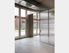 Innenansicht eines modernen Raumes mit Betonboden, großen weißen Fenstern und einer halbtransparenten Kunststofftrennwand mit Metallrahmen. Außerhalb der Fenster sind Backsteinfassaden und ein Gehweg sichtbar.