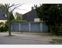 Reihe von vier grauen Garagentoren aus Metall, flankiert von Bäumen und Büschen in einer Wohngegend mit gepflastertem Bürgersteig davor. Im Hintergrund sind ein gelbes Wohnhaus sowie ein dunkel verkleidetes Gebäude sichtbar.