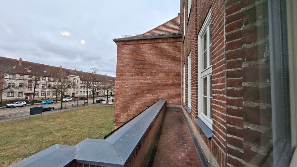 Blick von einem schmalen Balkon an einem Backsteingebäude mit weißen Sprossenfenstern und rotbraunem Fliesenboden. Im Hintergrund sind eine Straße mit parkenden Autos, eine Rasenfläche und gegenüberliegende Wohnhäuser unter bewölktem Himmel zu sehen.