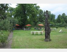 Grüne Parklandschaft mit Wiese, auf der sich eine hölzerne Skulptur befindet, die mehrere Figuren zeigt. Im Hintergrund stehen Tische und Stühle unter orangefarbenen Sonnenschirmen, umgeben von Bäumen und Sträuchern.